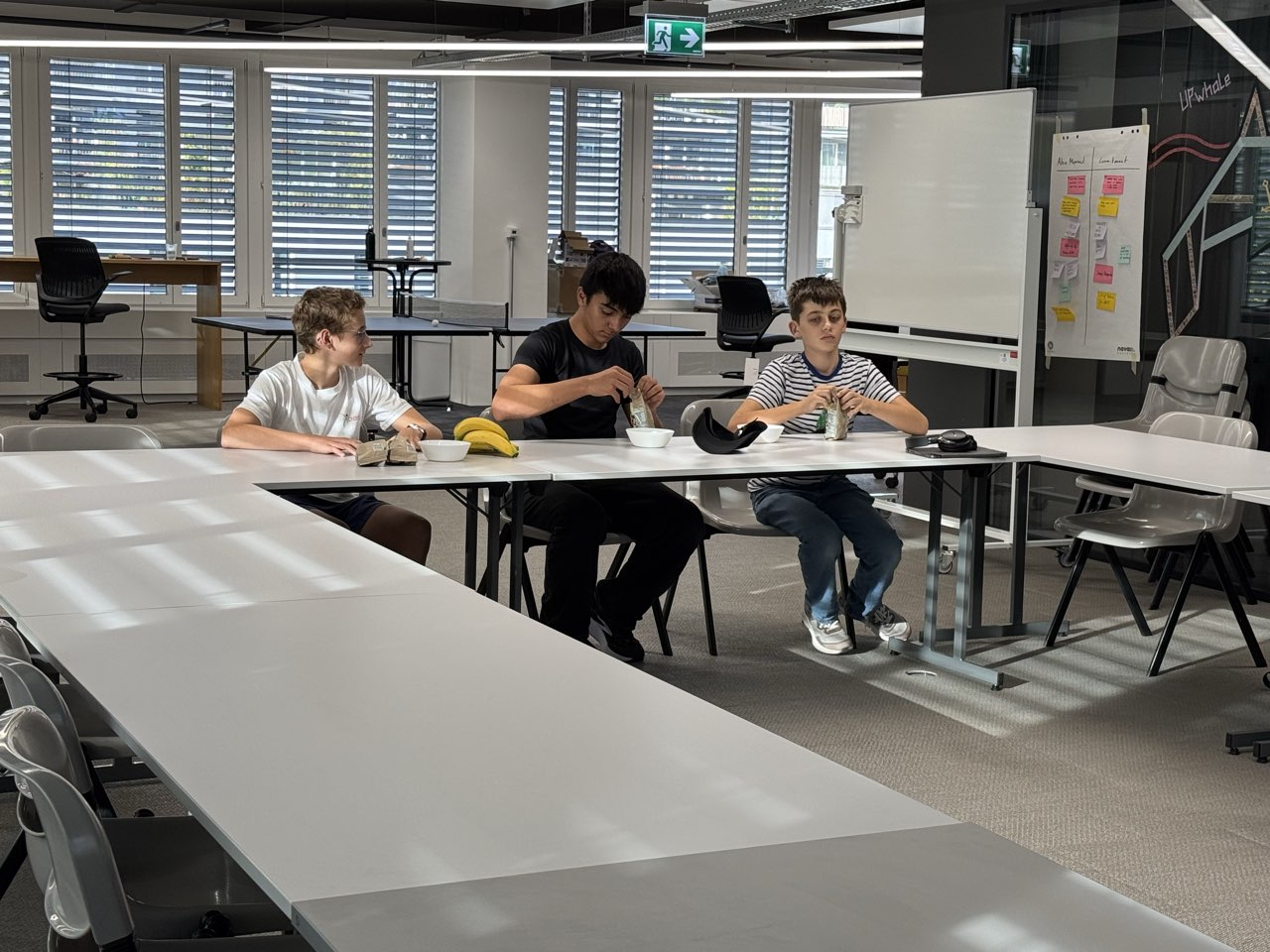 Drei Schüler sitzen an einem langen Tisch im Büro, dahinter ein Whiteboard mit Notizzetteln